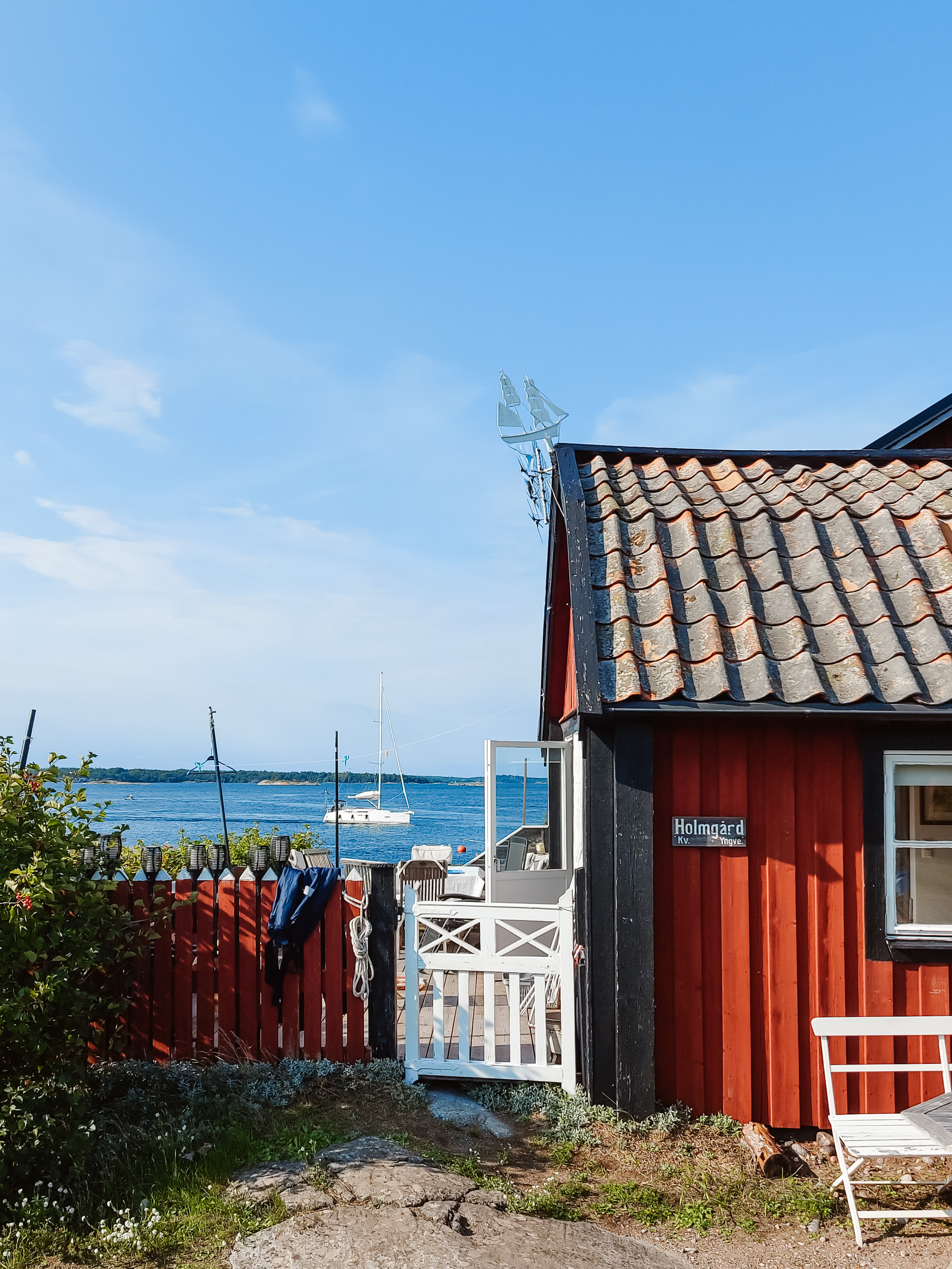 Fischerhütte aus rot gestrichenem Holz, dahinter fährt ein weißes Segelboot am Meer vorbei